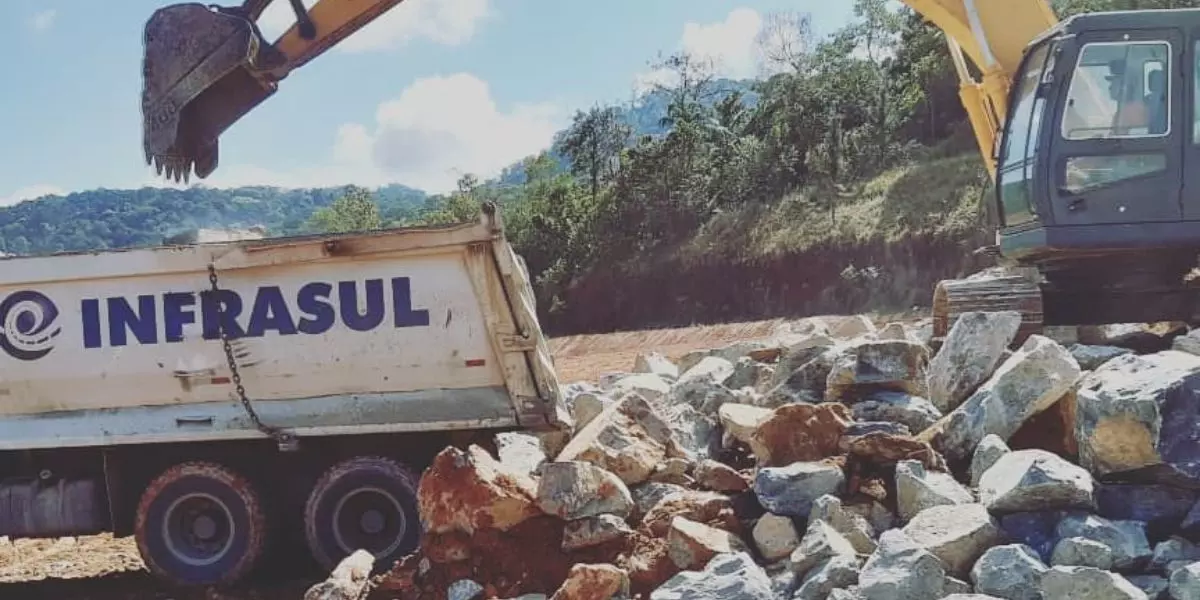 A foto mostra um caminhão basculante da infrasul sendo carregado por uma escavadeira Hidráulica