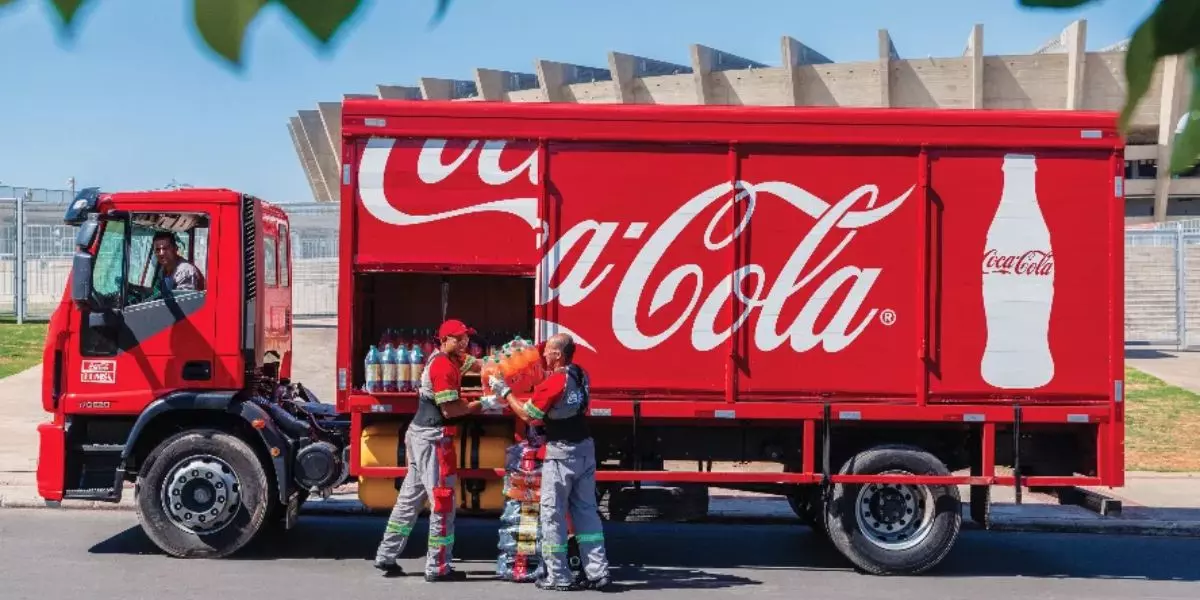 A foto mostra um caminhão da Coca-Cola e dois ajudantes e um motorista de caminhão